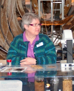 Judith Price stands behind display cases with specimens and a microscope on top, with blue-whale ribs in the background.