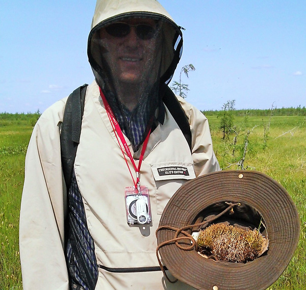 A man wearing bug netting holds a hat that contains a lichen specimen.