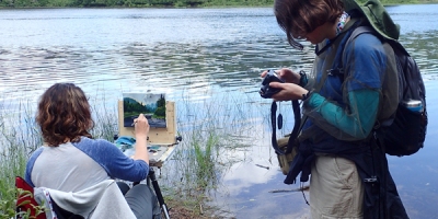A woman paints near the river, another woman examines photographs on her camera