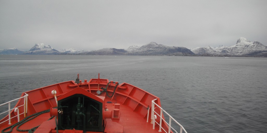 A ship’s bow in the foreground. Grey ocean and snow-capped mountains in the background.